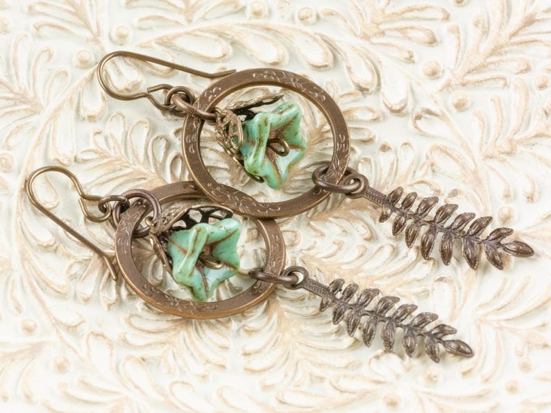 May include: A pair of handcrafted earrings with a bronze-coloured metal design. Each earring has a circular frame with a turquoise-coloured floral bead and a fern-shaped dangle. The earrings are displayed on a white, textured surface.