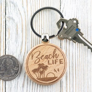 Pu&ograve; includere: Un portachiavi in legno con un'etichetta rotonda di colore marrone chiaro su cui &egrave; scritto "Beach Life" in caratteri bianchi. L'etichetta &egrave; incisa a laser con un'immagine di palme e un tramonto. Il portachiavi &egrave; attaccato a un anello portachiavi argentato.