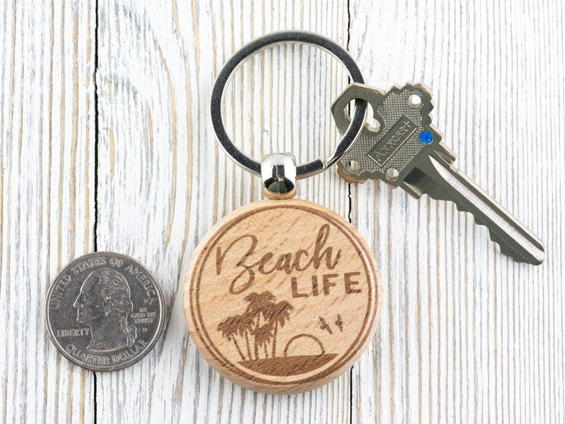 Pu&ograve; includere: Un portachiavi in legno con un'etichetta rotonda di colore marrone chiaro su cui &egrave; scritto "Beach Life" in caratteri bianchi. L'etichetta &egrave; incisa a laser con un'immagine di palme e un tramonto. Il portachiavi &egrave; attaccato a un anello portachiavi argentato.