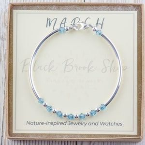 Puede incluir: Un brazalete de plata con un dije de cristal azul claro. El brazalete est&aacute; en una caja con el texto "MARCH" y "Black Brook Shop" en ella. La caja tambi&eacute;n dice "Joyer&iacute;a y relojes inspirados en la naturaleza".