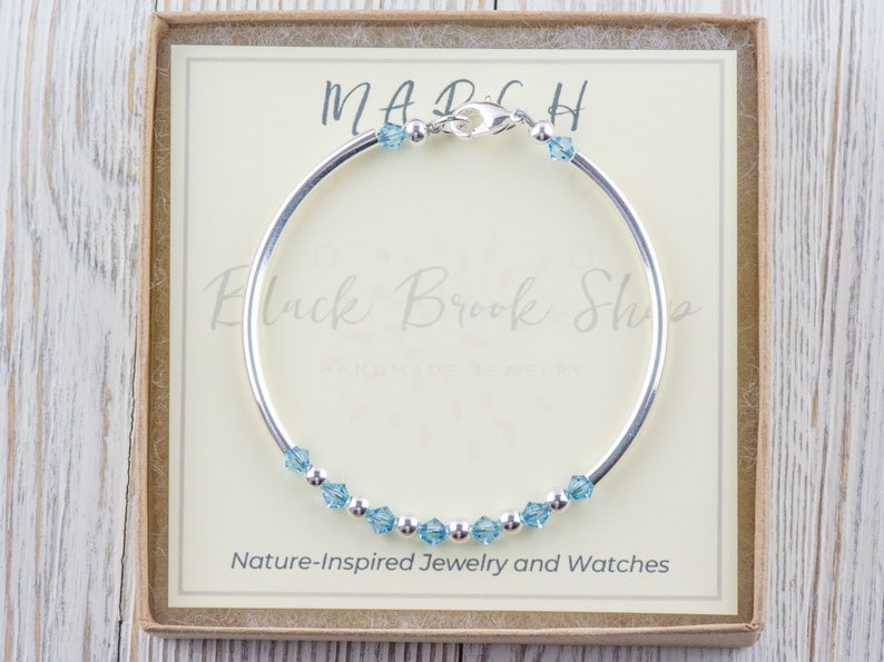 Puede incluir: Un brazalete de plata con un dije de cristal azul claro. El brazalete est&aacute; en una caja con el texto "MARCH" y "Black Brook Shop" en ella. La caja tambi&eacute;n dice "Joyer&iacute;a y relojes inspirados en la naturaleza".