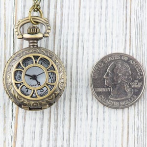Puede incluir: Un peque&ntilde;o reloj de bolsillo de estilo antiguo con una esfera blanca y agujas negras. El reloj est&aacute; en una cadena y es aproximadamente del tama&ntilde;o de una moneda de 25 centavos.
