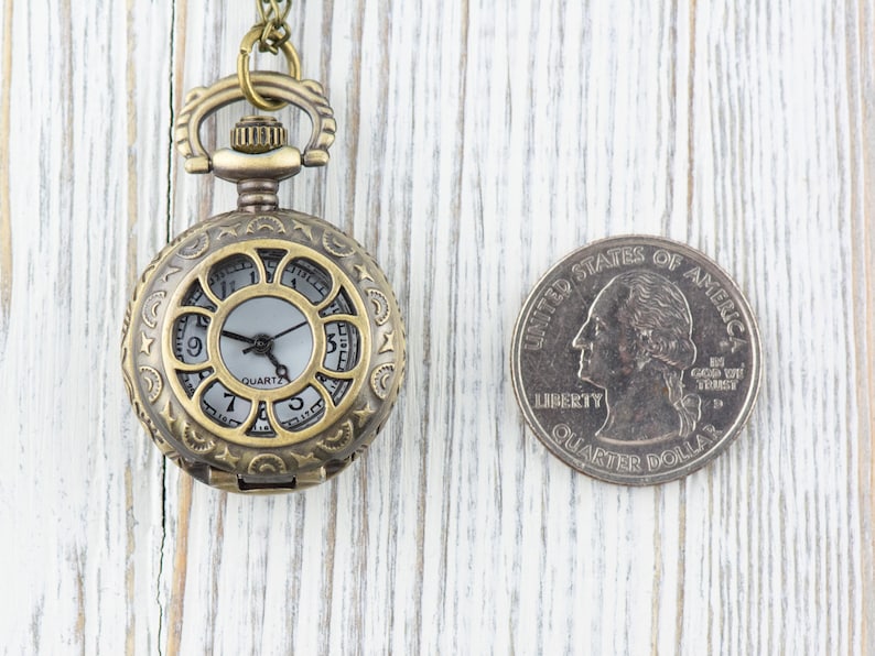 Puede incluir: Un peque&ntilde;o reloj de bolsillo de estilo antiguo con una esfera blanca y agujas negras. El reloj est&aacute; en una cadena y es aproximadamente del tama&ntilde;o de una moneda de 25 centavos.