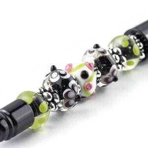 Op de afbeelding: Een zwarte pen met decoratieve glazen kralen. De kralen hebben verschillende ontwerpen, waaronder zwart-witte cirkels, roze accenten en groene stippen. De kralen worden afgewisseld met zilverkleurige metalen accenten.