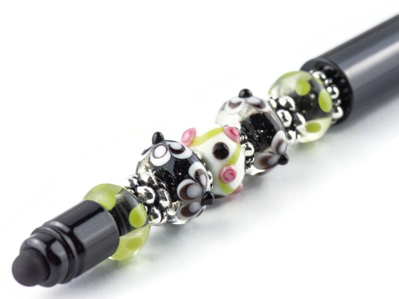 Op de afbeelding: Een zwarte pen met decoratieve glazen kralen. De kralen hebben verschillende ontwerpen, waaronder zwart-witte cirkels, roze accenten en groene stippen. De kralen worden afgewisseld met zilverkleurige metalen accenten.