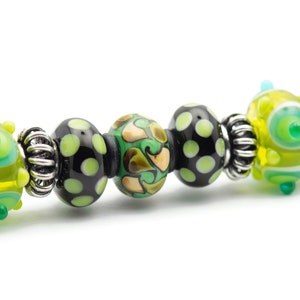 May include: A close-up of a silver charm bracelet with black, green, and yellow glass beads. The beads have various patterns, including polka dots, swirls, and abstract designs.