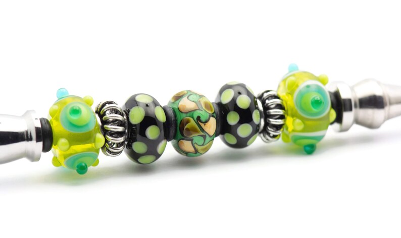 May include: A close-up of a silver charm bracelet with black, green, and yellow glass beads. The beads have various patterns, including polka dots, swirls, and abstract designs.