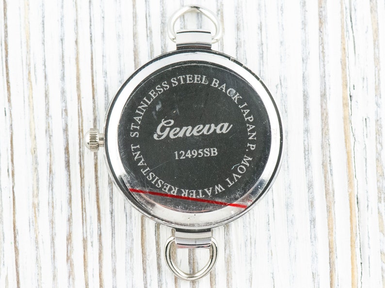 May include: The back of a silver watch with the text "STAINLESS STEEL BACK JAPAN MOV'T. WATER RESISTANT" and "Geneva 12495SB" engraved on it.