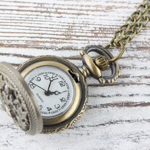 May include: A gold-toned pocket watch with a white face and black hands. The watch has a decorative, ornate design and is attached to a gold chain.