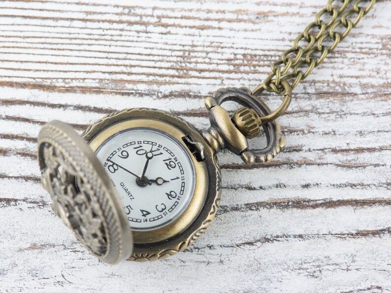 May include: A gold-toned pocket watch with a white face and black hands. The watch has a decorative, ornate design and is attached to a gold chain.