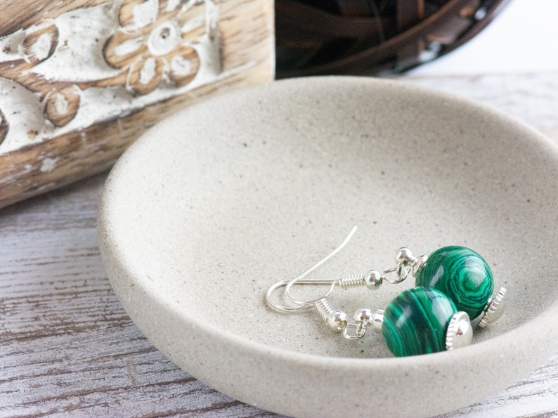 May include: A pair of silver earrings with green malachite stone beads. The earrings are on a white ceramic dish.