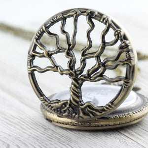 May include: Open antique bronze pocket watch featuring a tree of life design. The round watch has an openwork tree design. The watch is open, showing a white face. It rests on a light wood surface.