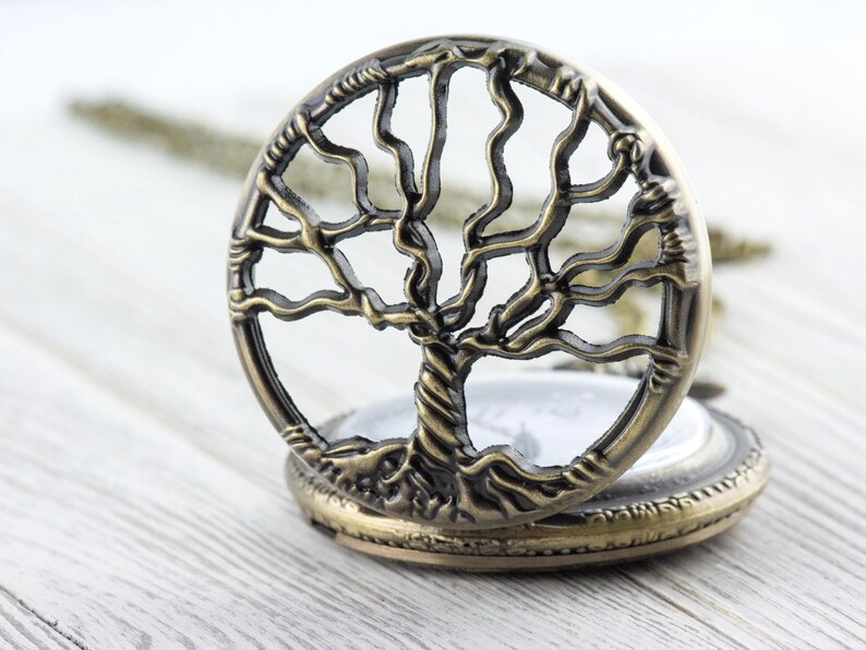 May include: Open antique bronze pocket watch featuring a tree of life design. The round watch has an openwork tree design. The watch is open, showing a white face. It rests on a light wood surface.