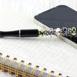 Op de afbeelding: Een zwarte pen met een decoratief kralengedeelte, liggend op een spiraalgebonden notitieboek en een smartphone. De pen heeft een zwarte schacht en kleurrijke kralen, waaronder groen, wit en zwart. Het notitieboek heeft een gouden spiraalbinding.