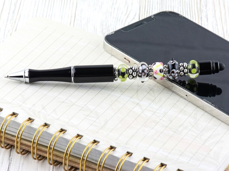 Op de afbeelding: Een zwarte pen met een decoratief kralengedeelte, liggend op een spiraalgebonden notitieboek en een smartphone. De pen heeft een zwarte schacht en kleurrijke kralen, waaronder groen, wit en zwart. Het notitieboek heeft een gouden spiraalbinding.