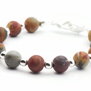 May include: A beaded bracelet featuring a series of round, multi-colored stone beads. The beads are primarily earth-toned, with shades of brown, gray, and orange. Small, silver-colored beads separate the larger stone beads. The bracelet is on a white background.