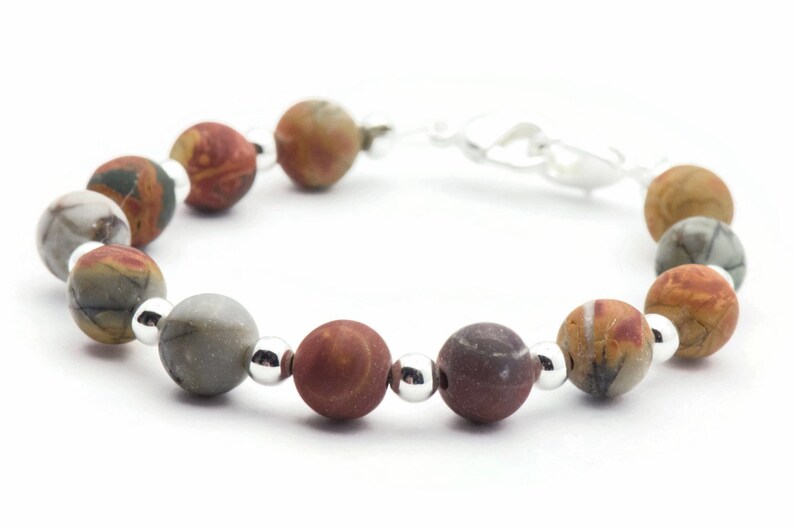 May include: A beaded bracelet featuring a series of round, multi-colored stone beads. The beads are primarily earth-toned, with shades of brown, gray, and orange. Small, silver-colored beads separate the larger stone beads. The bracelet is on a white background.