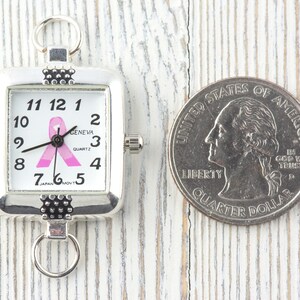 Pu&ograve; includere: Orologio quadrato color argento con quadrante bianco, numeri neri e design a nastro rosa. L'orologio &egrave; accanto a un quarto di dollaro USA. L'orologio ha un anello nella parte superiore e inferiore per il fissaggio.