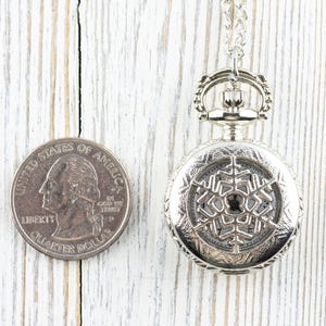 May include: A silver pocket watch with a snowflake design on the face. The watch is about the size of a UK 20 pence coin.