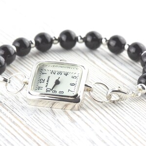 Puede incluir: Un reloj de esfera cuadrada con esfera blanca y agujas negras est&aacute; unido a una pulsera de grandes cuentas negras redondas. El reloj tiene una caja plateada y un cierre plateado. La pulsera se exhibe sobre una superficie de madera clara.