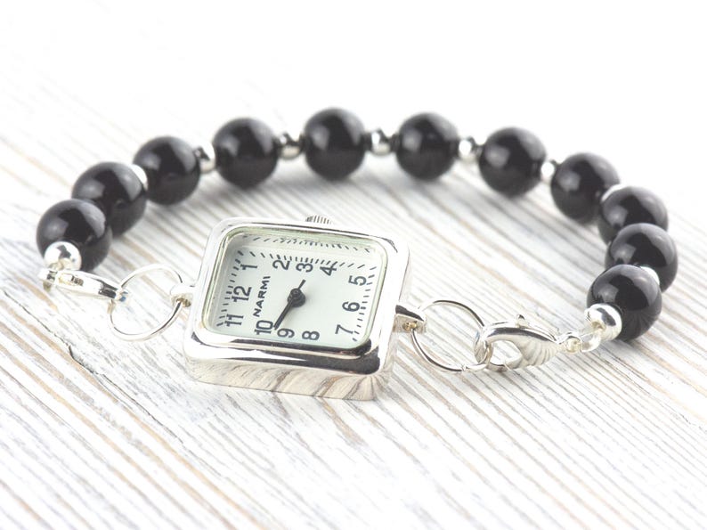 Puede incluir: Un reloj de esfera cuadrada con esfera blanca y agujas negras est&aacute; unido a una pulsera de grandes cuentas negras redondas. El reloj tiene una caja plateada y un cierre plateado. La pulsera se exhibe sobre una superficie de madera clara.