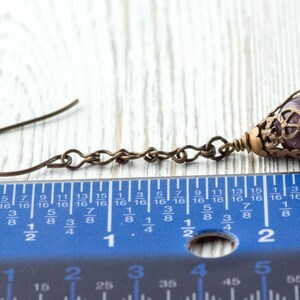 May include: A single antique bronze earring with a purple gemstone bead. The earring features a decorative filigree cap and a chain link design. The earring is approximately 2.5 cm long, as shown by the ruler.