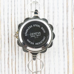 May include: The back of a silver watch with a round, scalloped edge. The watch face says "Stainless Steel Back", "Geneva", "Japan Movt C-11", and "Water Resistant".