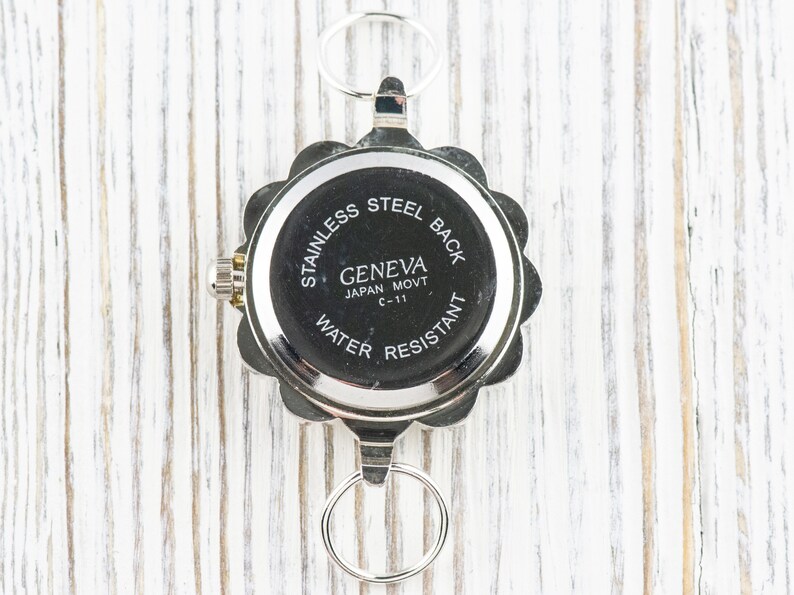 May include: The back of a silver watch with a round, scalloped edge. The watch face says "Stainless Steel Back", "Geneva", "Japan Movt C-11", and "Water Resistant".