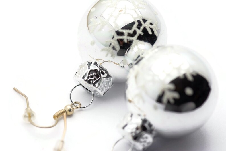 May include: Silver Christmas ornament earrings with snowflake designs. The earrings have a silver hook and a silver bead.
