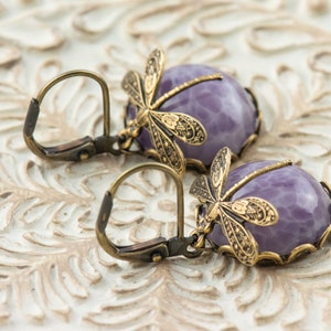 Pode incluir: Um par de brincos pendentes com caboch&otilde;es de ametista e detalhes de lib&eacute;lula. Os brincos s&atilde;o feitos de lat&atilde;o e t&ecirc;m um estilo vintage.