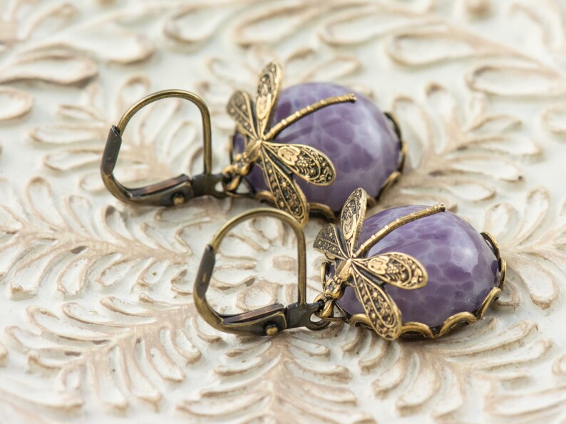 Pode incluir: Um par de brincos pendentes com caboch&otilde;es de ametista e detalhes de lib&eacute;lula. Os brincos s&atilde;o feitos de lat&atilde;o e t&ecirc;m um estilo vintage.