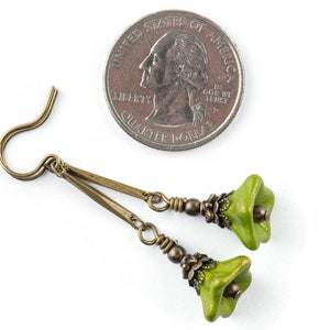 Puede incluir: Un par de pendientes de color verde en forma de flor con detalles met&aacute;licos de color bronce y ganchos. Los pendientes se muestran junto a una moneda de veinticinco centavos de d&oacute;lar estadounidense para comparar el tama&ntilde;o.