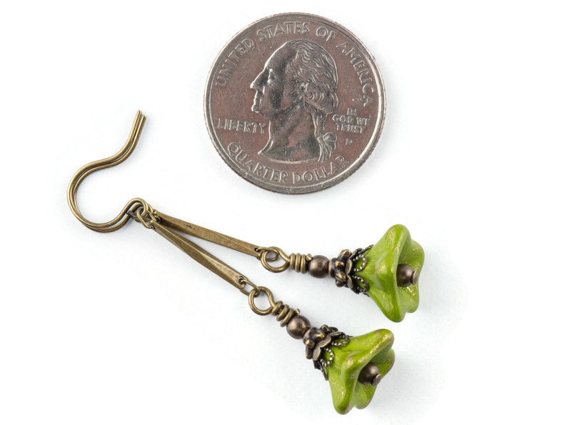 Puede incluir: Un par de pendientes de color verde en forma de flor con detalles met&aacute;licos de color bronce y ganchos. Los pendientes se muestran junto a una moneda de veinticinco centavos de d&oacute;lar estadounidense para comparar el tama&ntilde;o.