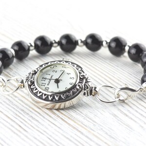 Op de afbeelding: Een zilverkleurige horlogearmband met een witte wijzerplaat en een zwarte kralenband. De wijzerplaat heeft zwarte cijfers en wijzers. De armband is gemaakt van gepolijste zwarte kralen en zilverkleurige metalen schakels en sluiting.