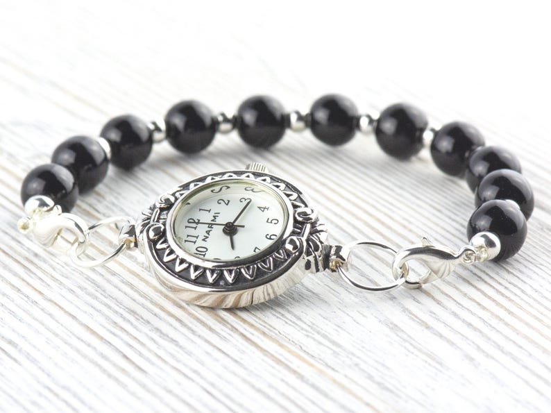 Op de afbeelding: Een zilverkleurige horlogearmband met een witte wijzerplaat en een zwarte kralenband. De wijzerplaat heeft zwarte cijfers en wijzers. De armband is gemaakt van gepolijste zwarte kralen en zilverkleurige metalen schakels en sluiting.