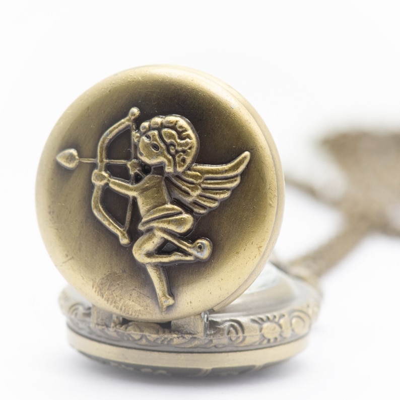 Puede incluir: Un reloj de bolsillo dorado con un dise&ntilde;o ornamentado. La tapa presenta una escultura en relieve de un querub&iacute;n alado sosteniendo un arco y una flecha.