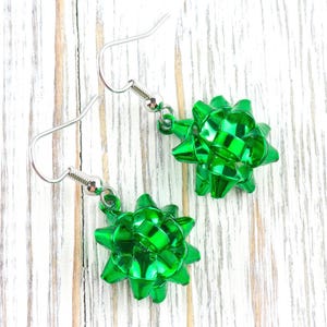 May include: A pair of green bow-shaped earrings with silver-colored fish hook ear wires. The earrings are made of shiny, metallic material and are set against a white wooden background.