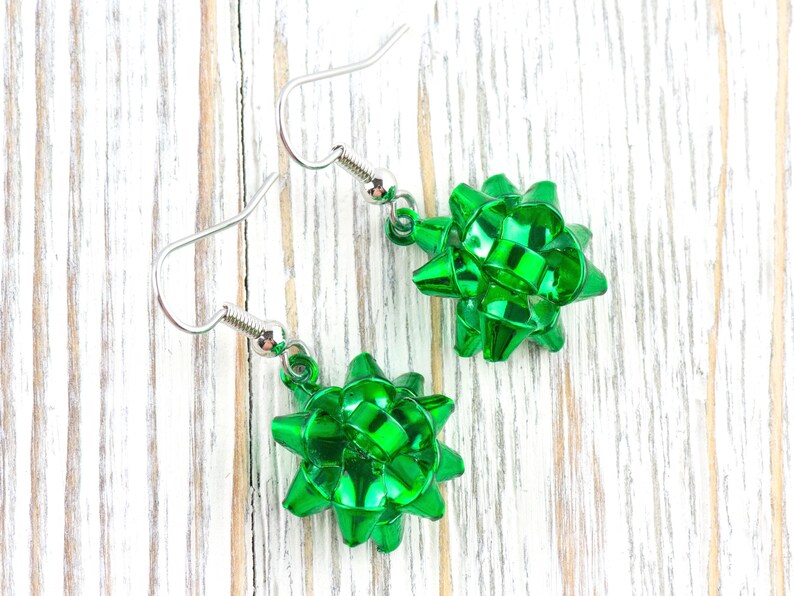 May include: A pair of green bow-shaped earrings with silver-colored fish hook ear wires. The earrings are made of shiny, metallic material and are set against a white wooden background.