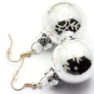 May include: Silver Christmas ornament earrings with snowflake designs. The earrings are attached to gold hooks.