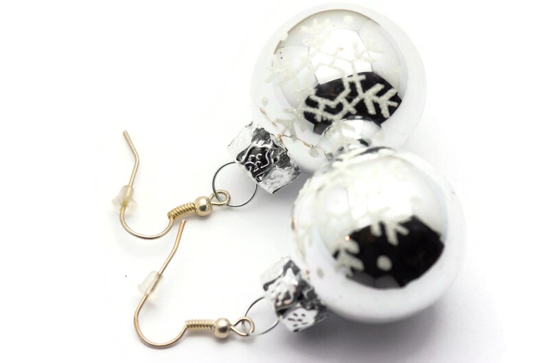 May include: Silver Christmas ornament earrings with snowflake designs. The earrings are attached to gold hooks.