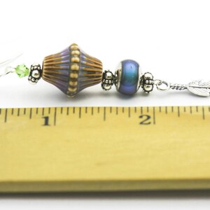 Pu&ograve; includere: Un orecchino d'argento con un ciondolo a forma di piuma argentata, una perla blu iridescente e una perla con un motivo viola, marrone e oro. L'orecchino &egrave; lungo circa 5 cm.