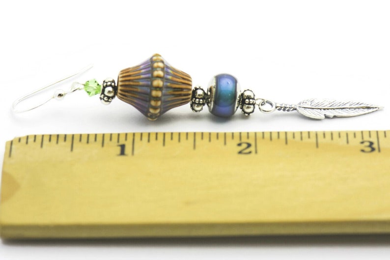 Pu&ograve; includere: Un orecchino d'argento con un ciondolo a forma di piuma argentata, una perla blu iridescente e una perla con un motivo viola, marrone e oro. L'orecchino &egrave; lungo circa 5 cm.