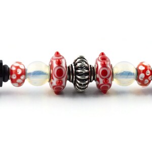 Puede incluir: Primer plano de una pulsera con cuentas de vidrio rojas y blancas, una cuenta espaciadora plateada y tapas finales negras.