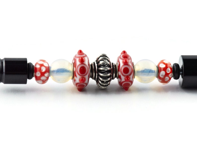 Puede incluir: Primer plano de una pulsera con cuentas de vidrio rojas y blancas, una cuenta espaciadora plateada y tapas finales negras.