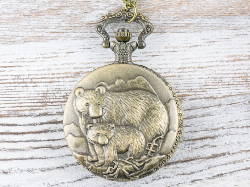May include: A gold-toned pocket watch with a detailed design of a mother bear and her cub. The watch has a decorative chain attached.
