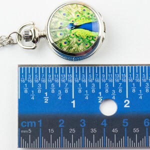 May include: A silver pocket watch with a chain. The watch face features a colourful peacock design. The watch is 2.5 cm in diameter.