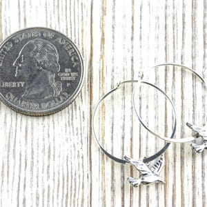 Peut inclure: Boucles d'oreilles cr&eacute;oles en argent avec un charm en forme d'oiseau. Les boucles d'oreilles sont pr&eacute;sent&eacute;es &agrave; c&ocirc;t&eacute; d'un quart de dollar am&eacute;ricain pour comparaison de taille. Sur une surface en bois blanc.