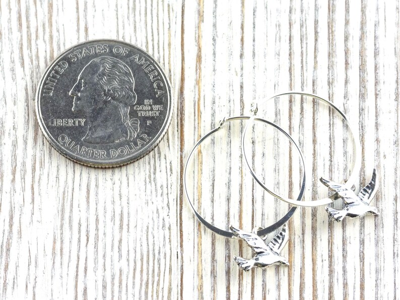 Peut inclure: Boucles d'oreilles cr&eacute;oles en argent avec un charm en forme d'oiseau. Les boucles d'oreilles sont pr&eacute;sent&eacute;es &agrave; c&ocirc;t&eacute; d'un quart de dollar am&eacute;ricain pour comparaison de taille. Sur une surface en bois blanc.