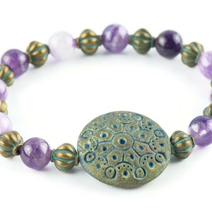 May include: A purple amethyst gemstone bracelet with a teal blue and bronze patterned charm. The bracelet is made with round beads and has a rustic, bohemian style.