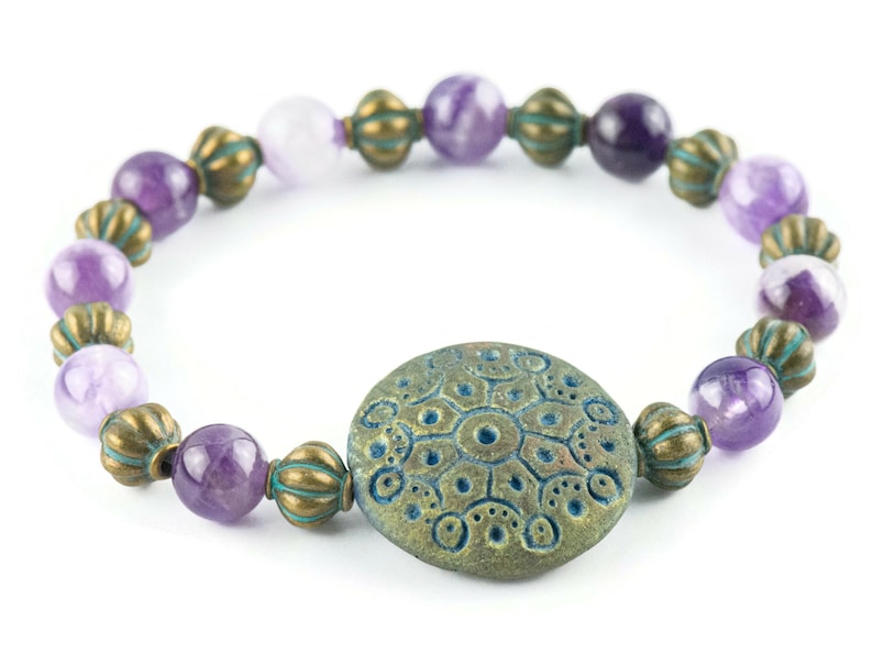 May include: A purple amethyst gemstone bracelet with a teal blue and bronze patterned charm. The bracelet is made with round beads and has a rustic, bohemian style.